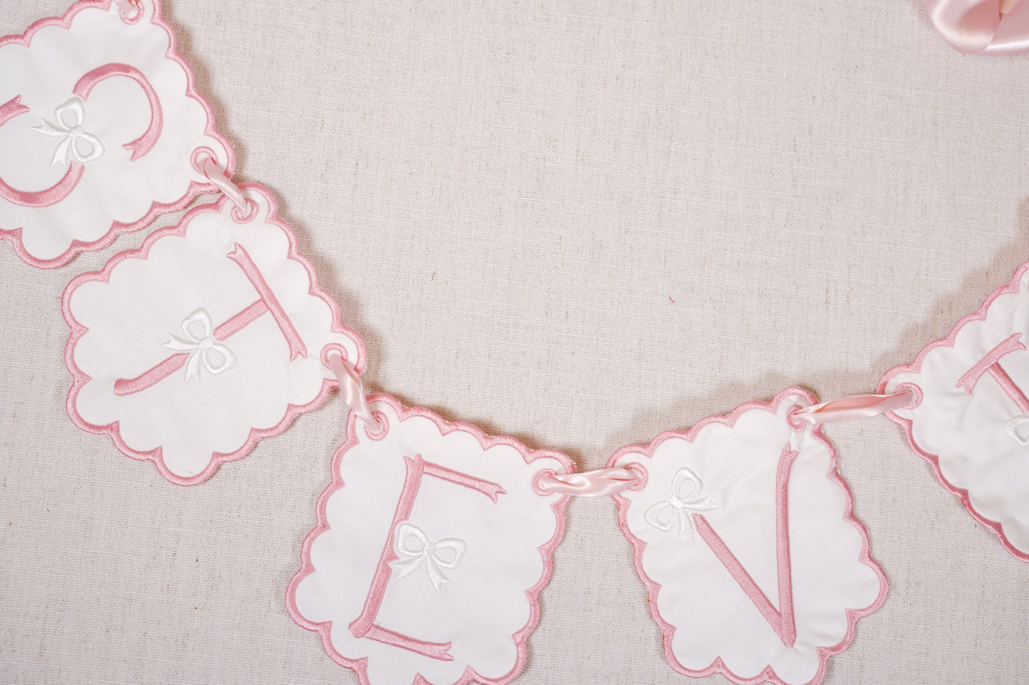 Custom embroidered baby name garland spelling stevie with scalloped pink edges and satin ribbon, displayed on neutral linen backdrop.