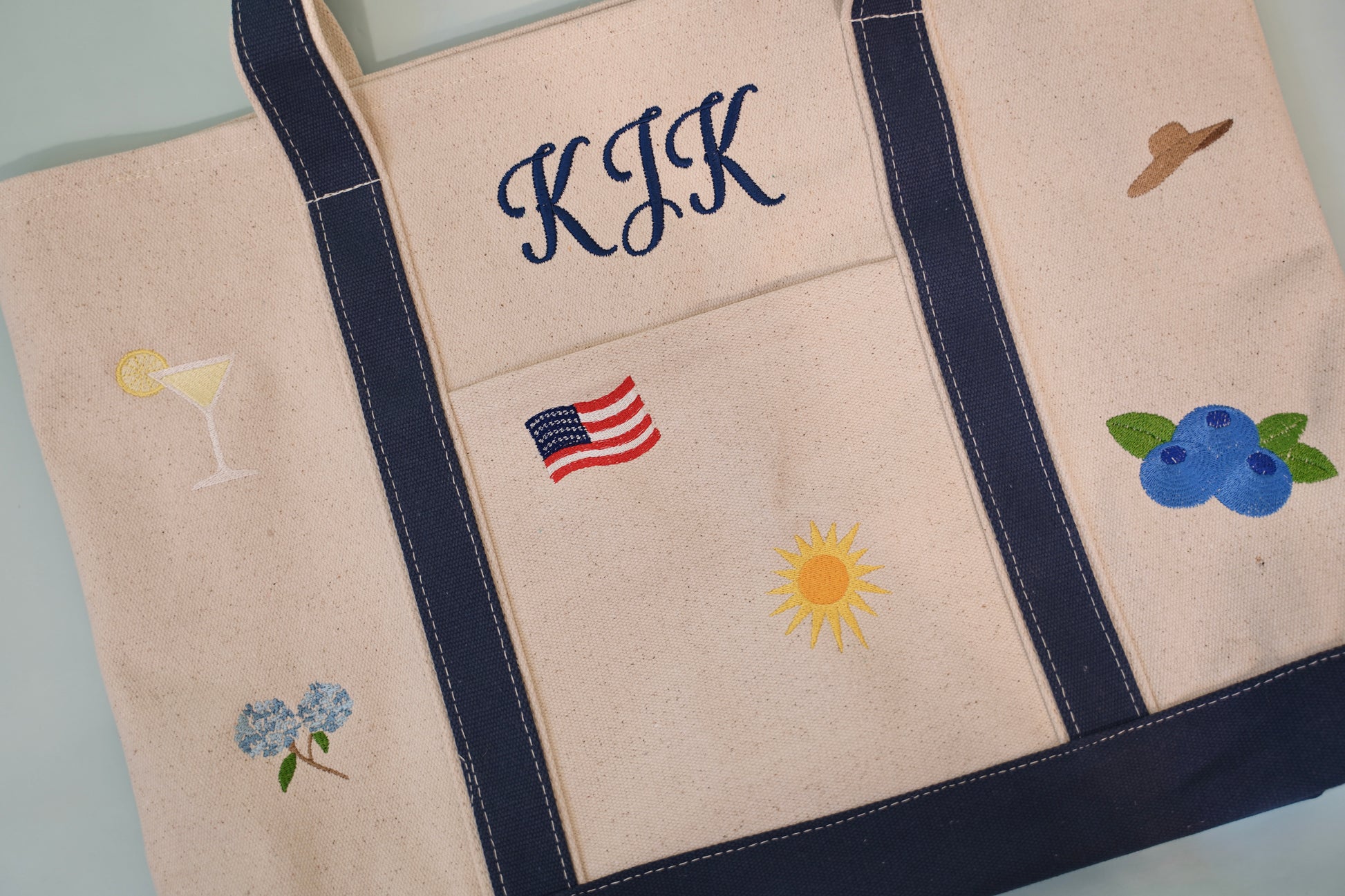 Close-up detail of embroidered hydrangea, sun, blueberries, American flag, and sun hat on canvas tote bag

