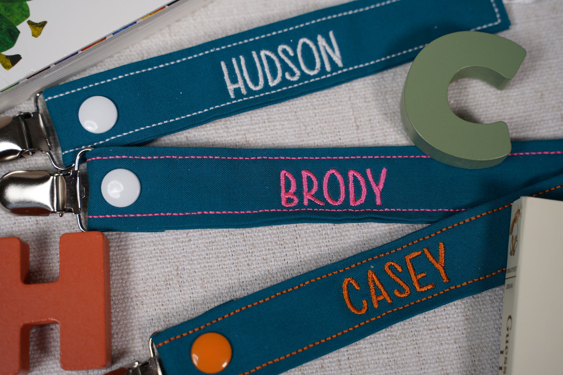 “Set of three personalized embroidered pacifier clips in teal fabric, featuring names ‘Hudson’ in white, ‘Brody’ in pink, and ‘Casey’ in orange. Each clip has contrasting stitching, secure metal clips, and snap button closures. Displayed with wooden letter blocks and a children’s book on a neutral fabric background.”