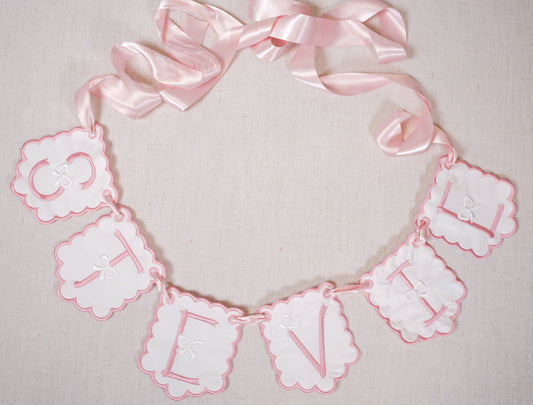 Custom embroidered baby name banner with pink scalloped pendants and satin ribbon, spelling “stevie”; heirloom nursery décor by Kits Threads.